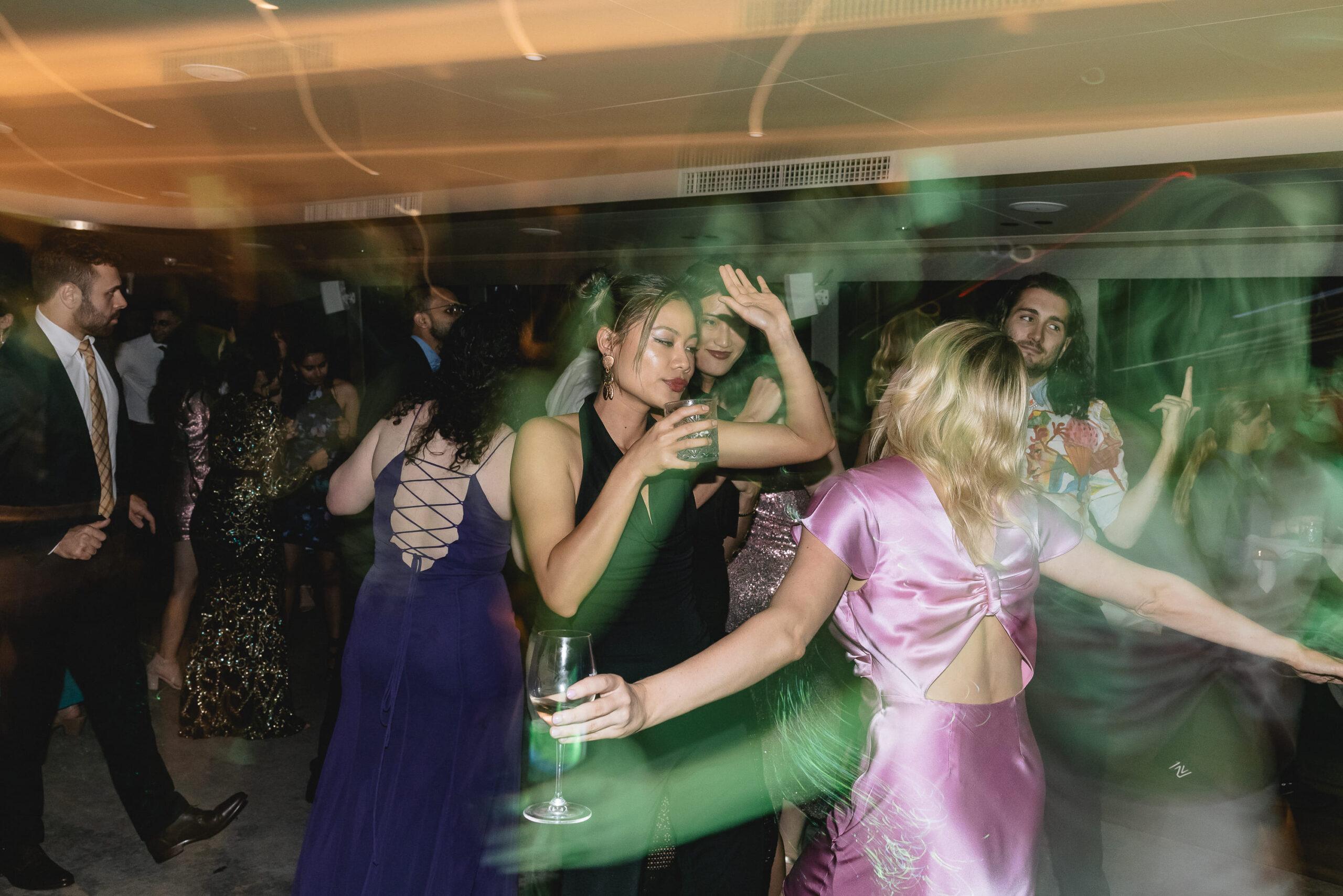 People in formal evening attire dance and hold drinks at a dimly lit indoor party with motion blur light effects.
