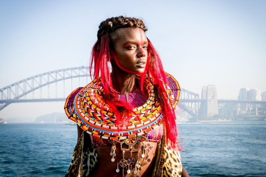 Young woman with bright red hair and ornate beaded collar posing in front of the Sydney Harbour Bridge.