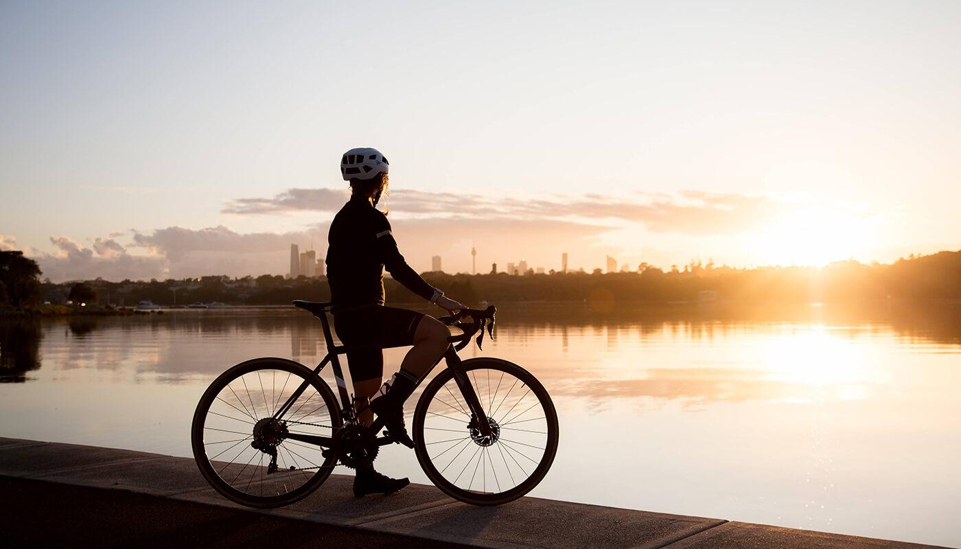 Cyclist in helmet stands with a bicycle by the water during a golden sunset with a city skyline in the distance.