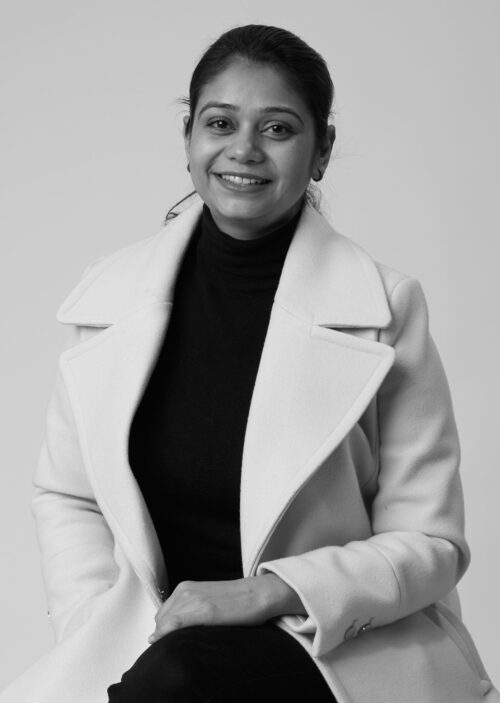 Smiling woman with pulled-back hair wearing a black turtleneck under a light-colored coat against a plain background.