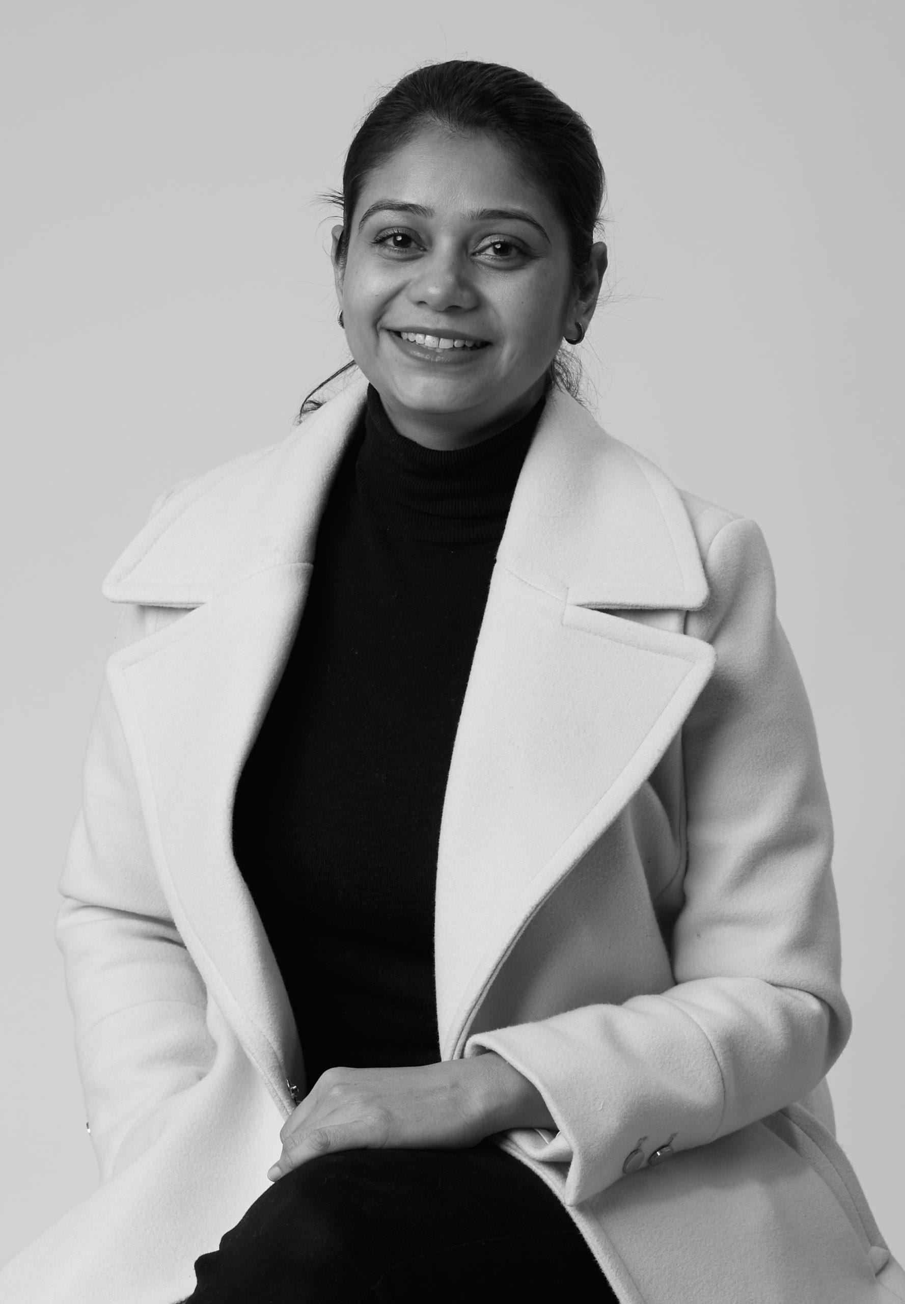 Smiling woman with pulled-back hair wearing a black turtleneck under a light-colored coat against a plain background.