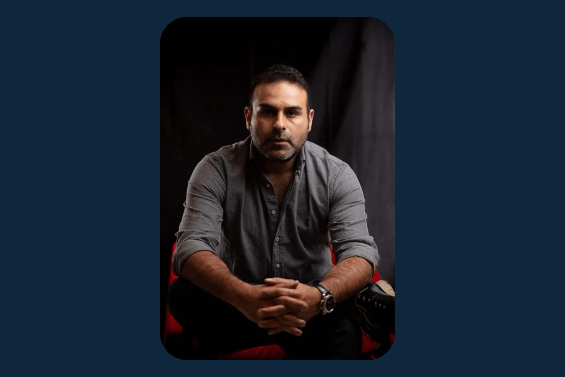 Man with short dark hair and a beard wearing a grey button-down shirt sitting against a dark background.