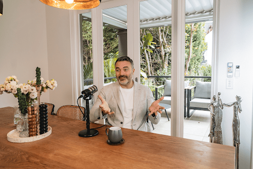 Man in a light blazer gestures while speaking into a microphone at a wooden dining table.